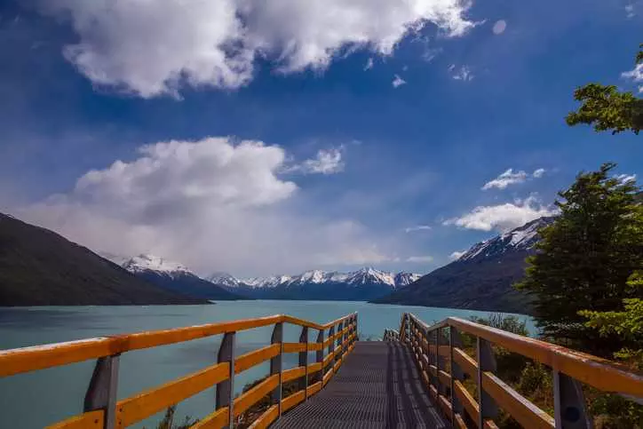 Пейзажный вид на озеро Аргентины (Lago Argentino), заснеженные горы сзади, туристическая лестница спереди. Голубое небо с красивыми облаками